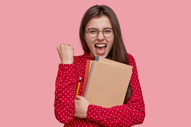 Positive happy teacher clenches fist with joyful expression, holds spiral notepad, writes notes in notepad, dressed in red shirt