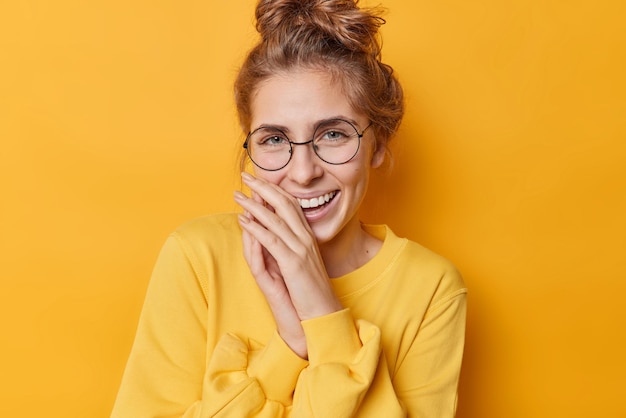 Positive happy woman laughs gladfully keeps hands near mouth expresses authentic sincere emotions wears spectacles and casual jumper isolated over vivid yellow background Happiness concept