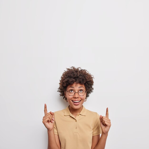 Positive happy woman with curly hair points above shows nice advertisement overhead dressed in casual beige t shirt shows promo or nice discount isolated over beige background. Wow look there