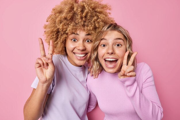 Positive happy women have fun carefree mood make peace gesture or v sign have glad expressions stand closely dressed in casual clothes isolated over pink background express optimism and joy
