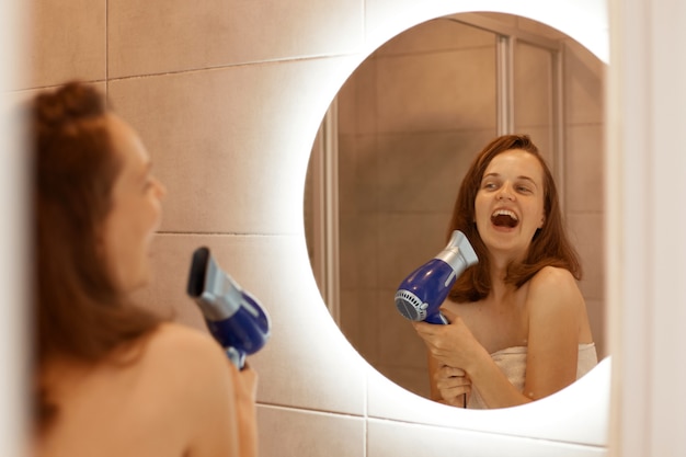 Positiva felice giovane donna adulta che asciuga i capelli in bagno con asciugacapelli, guardando il riflesso nello specchio e cantando, tiene la bocca aperta, procedure mattutine.