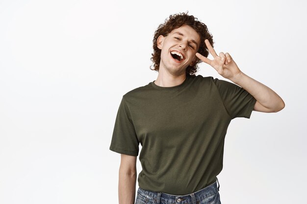 Positive happy young guy showing peace salute gesture and laughing concept of joy and happiness standing over white background