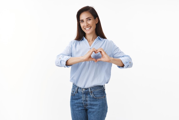 Positivo sano felice donna di mezza età mostra il segno del cuore sorridente amare la relazione esprimere amore cura e gioia confessare sentimenti per la simpatia della famiglia stand con ammirazione sfondo bianco