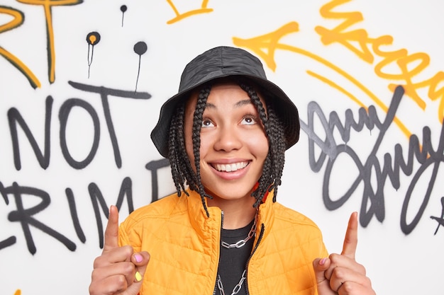 Positive hipster girl with braids smiles broadly points index fingers overhead dressed in fashionable clothes demonstrates something against graffiti wall belongs to youth subculture