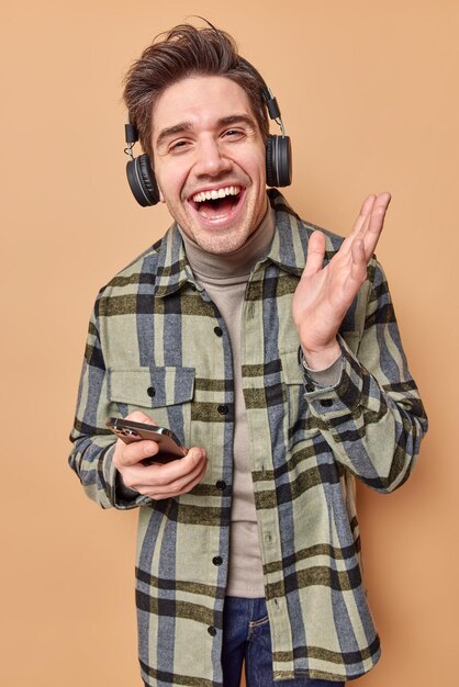 Positive hipster guy with dark hair wears checkered shirt listens music via headphones holds mobile phone enjoys favorite favorite song from playlist isolated on beige has happy mood