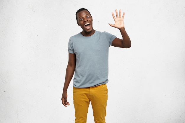 Free photo positive human emotions, facial expressions, feelings, attitude and reaction. friendly-looking polite young african american man dressed in grey t-shirt and mustard jeans saying hi, waving his hand