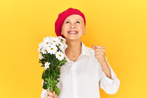 Positive human emotions, feelings and reaction. emotional ecstatic retired female in elegant head-wear and white shirt looking up and smiling, holding daisies, clenching fist, excited with success