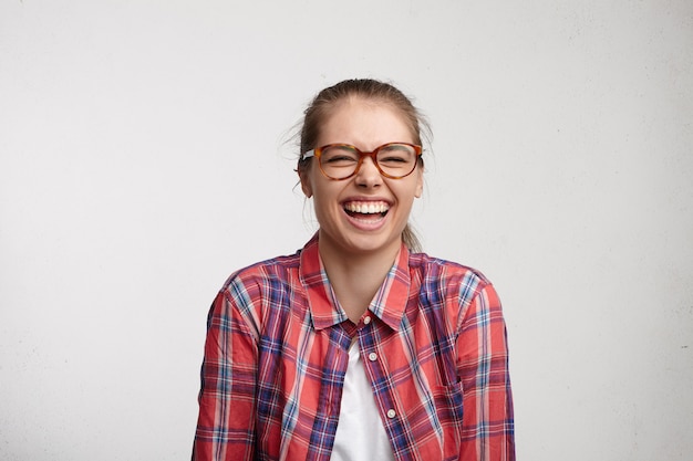 Positive human emotions. Studio portrait of relaxed carefree young woman with toothy smile wearing eyeglasses closing eyes tight while laughing out loud at good jokes, having fun with friends indoors