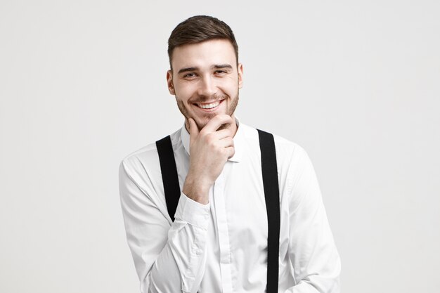 Positive human facial expression, emotions, feelings and life perception. Joyful handsome young bearded guy dressed in stylish white shirt with suspenders, laughing at joke, rubbing his stubble