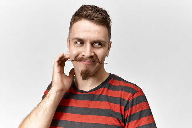 Positive human facial expressions, emotions and feelings. Close up shot of handsome charismatic hipster guy with goatee and curled mustache touching face and looking away with charming smile