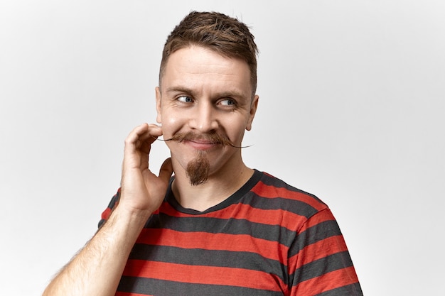 Positive human facial expressions, emotions and feelings. Close up shot of handsome charismatic hipster guy with goatee and curled mustache touching face and looking away with charming smile