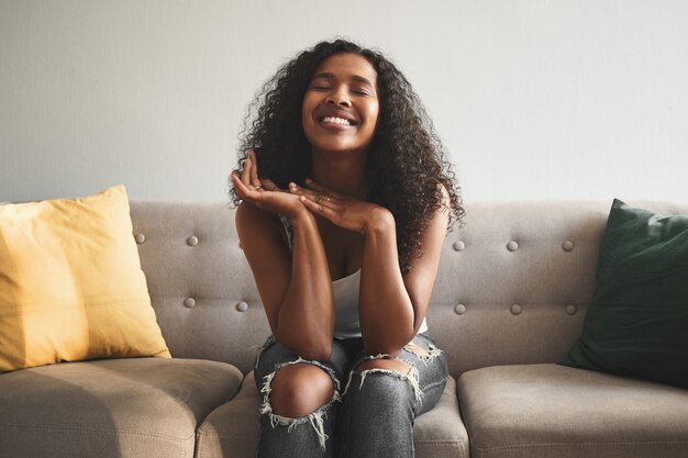 Positive human facial expressions, emotions, feelings and reaction. Indoor shot of emotional happy young mixed race woman wearing ripped jeans, closing eyes and smiling broadly, expressing joy