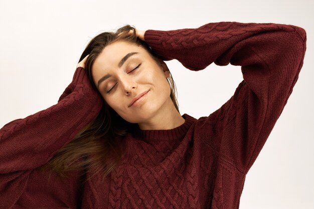 Positive human facial expressions and emotions. Isolated shot of charming young Caucasian female in knitted sweater keeping eyes closed with enjoyment, massaging head and smiling joyfully