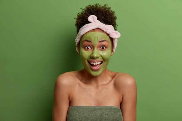 Positive joyful woman applies nutrient green mask on face, has daily hygienic routine, does morning beauty procedures, laughs happily, has healthy glowing skin, wrapped in bath towel, stands indoor