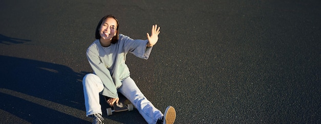 Бесплатное фото Позитивная корейская девушка закрывает лицо от солнечного света, сидит на скейтборде и счастливо улыбается.