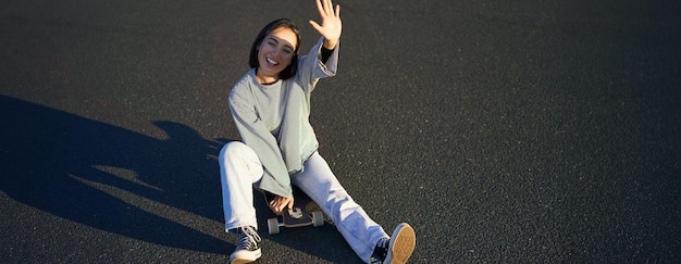 Foto gratuita una ragazza coreana positiva si copre il viso dalla luce del sole si siede sullo skateboard e sorride felicemente