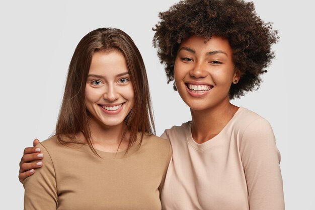 Positive ladies of different races stand next to each other, have warm hug, pleasant smile, friendly relationships