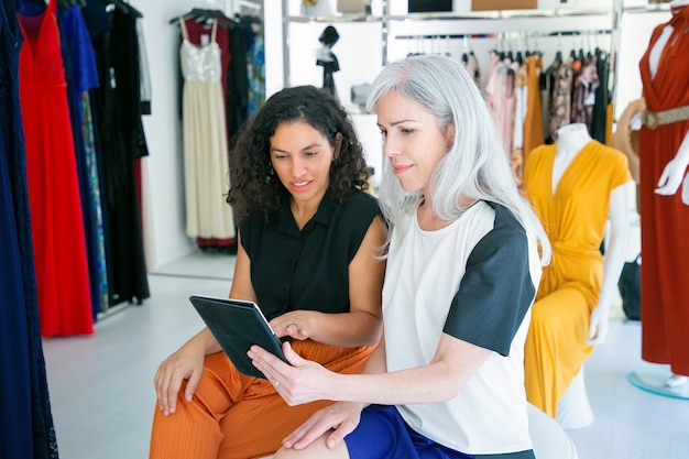 Позитивные дамы сидят вместе и используют планшет, обсуждают одежду и покупки в магазине модной одежды. Потребительство или концепция покупок