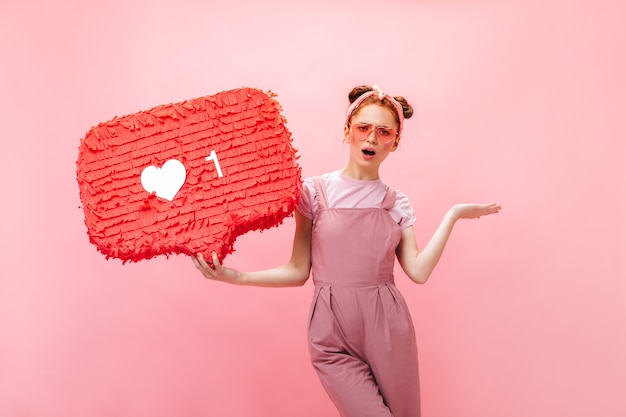 Free photo positive lady dressed in pink overalls, t-shirt and pink accessories points finger at like sign.