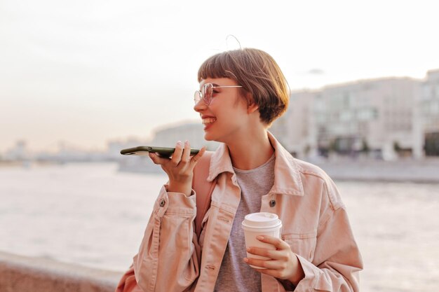 川の背景に電話を保持しているポジティブな女性ライトジャケットとメガネのショートヘアの女性が外でお茶を飲みながらポーズをとる
