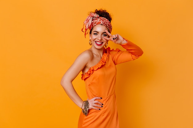 Positive lady in orange elegant dress and fashionable headband sincerely smiles and shows peace sign on orange space.