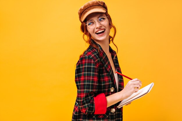 Positive lady in plaid outfit and cap poses with notebook on isolated wall