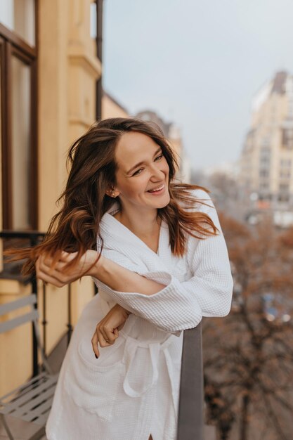 Positive lady in white robe touches her hair and laughs enjoying autumn day on her terrace