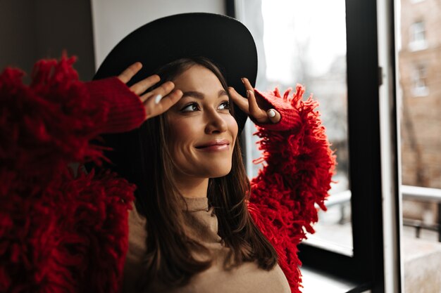 Positive lady with gloss on her lips in good mood looks out window. Picture of woman in red eco coat and hat.