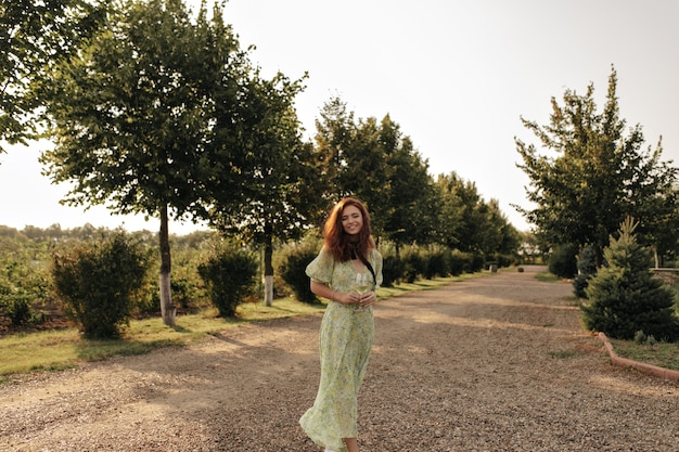 Positive lady with red hair and black bandage on her neck in long green dress smiling and holding glass with champagne on wall of road