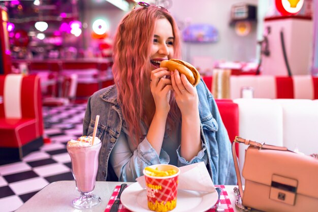 Портрет позитивного образа жизни счастливой вышедшей красивой женщины с розовыми волосами, обедающей в винтажном американском кафе, едящей хот-дог, картофелем фри и коктейлем, фастфудом, читмилом, пастельными тонами.