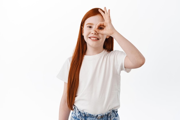 Positive little girl child with long red hair and freckles shows okay sign on eye and smile make OK gesture to praise and recommend good thing white background