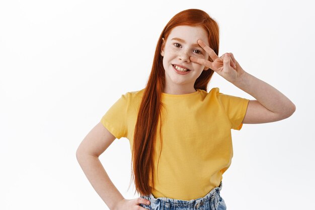 Positive little girl with long ginger hair and freckles smiling happy, showing peace kawaii sign on eye, posing in yellow t-shirt against white background