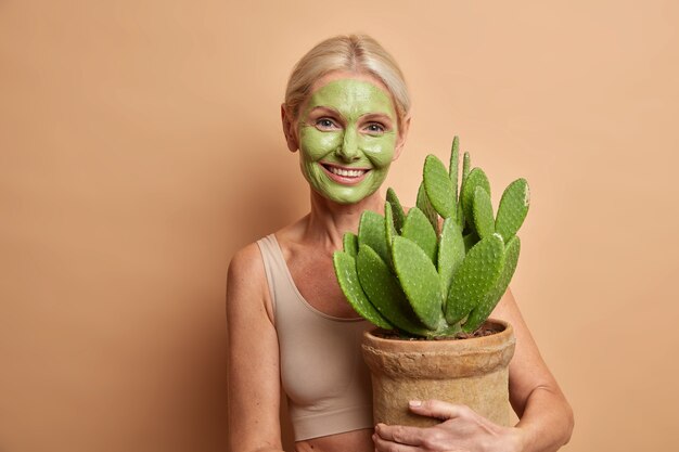 Positive lovely middle aged woman cares about skin applies green nourishing mask on face embraces pot with cactus smiles gently isolated over brown wall