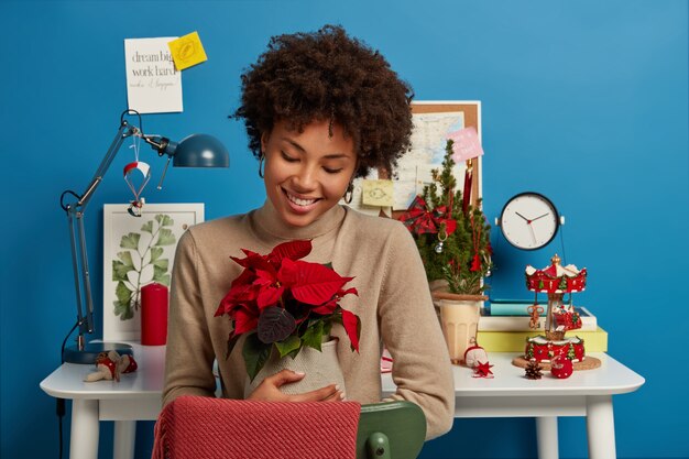 Positive lovely woman embraces vase with beautiful red flower, being in high spirit, smiles gently, enjoys cozy domestic atmosphere in cabinet