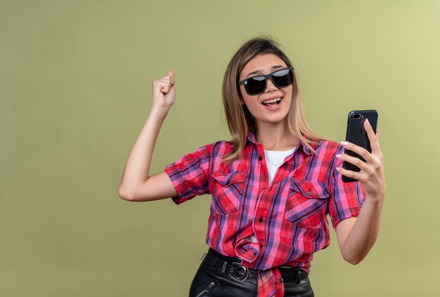 A positive lovely young woman in a checked shirt taking selfie with mobile phone in sunglasses while raising clenched fist on a green wall