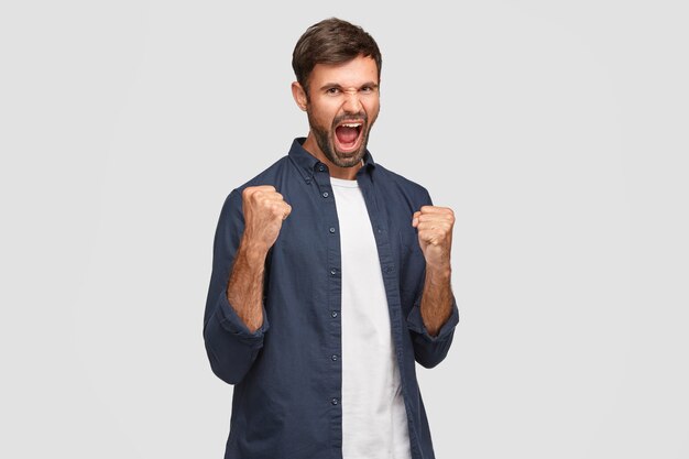 Positive male champion with happy expression, feels overjoyed as achieved success and won contest, exclaims with widely opend mouth, clenches fists, dressed in fashionable clothes, isolated on white