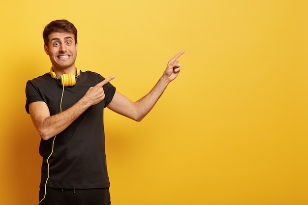 Positive male model clenches teeth, points at blank space for your text or advertisement, wears headphones on neck