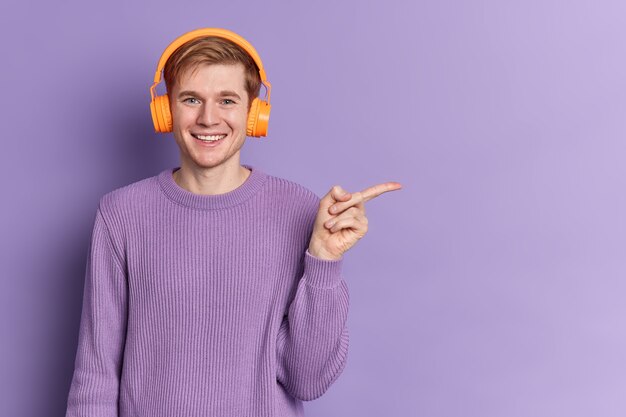 Позитивный подросток-мужчина с голубыми глазами и счастливой улыбкой в повседневном фиолетовом свитере слушает музыку в стереонаушниках. Молодежное хобби