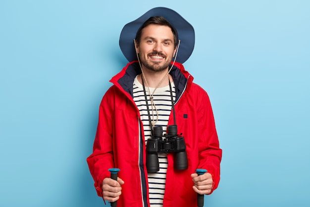 Positive male traveler uses trekking sticks for walking in forest, spends vacation actively, smiles positively, dressed in stylish headgear and red jacket, has binoculars on neck