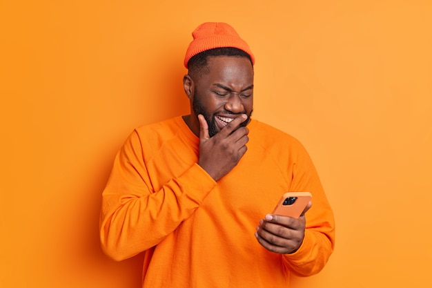 スマートフォンの画面に積極的に集中して笑うポジティブな男は、インターネットで面白いビデオを見たり、オレンジ色の壁に隔離されたカジュアルな明るい服を着た受信メッセージを笑ったりします