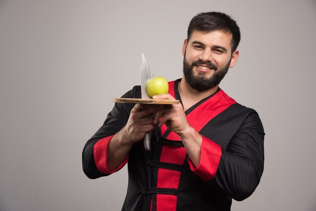 Positive man holding an apple with knife on wooden board .