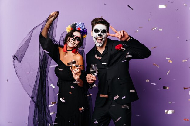 Positive man and lady in black clothes and masquerade masks sincerely rejoice and laugh, dancing among confetti at Halloween party.