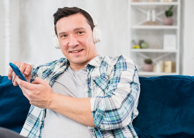 Positive man listening music in headphones and browsing on smartphone on sofa
