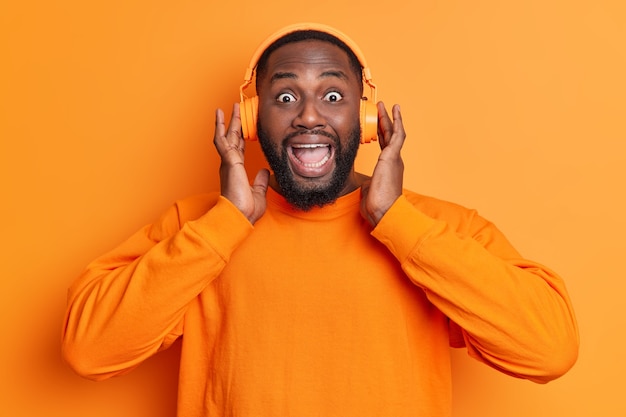 Positive man looks surpisingly at front being entertained listens favorite music via stereo headphones surprised by something wears long sleeved jumper isolated over orange wall