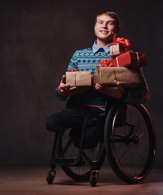 어두운 회색 배경 위에 크리스마스 선물을 들고 휠체어를 탄 긍정적인 남자.