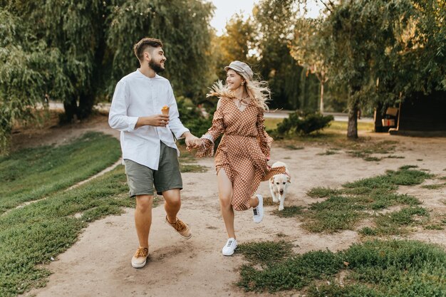Positive man with ice cream is holding hand of smiling woman in brown dress Romantic couple walking with big labrador in park