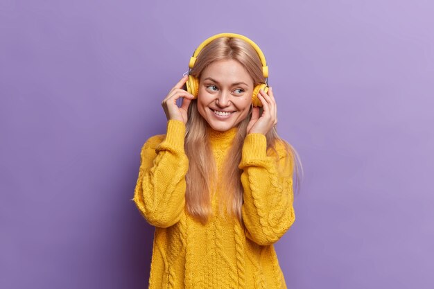 Positive millennial girl enjoys pleasant music via headphones being in good mood smiles happily dressed in kitted yellow jumper 