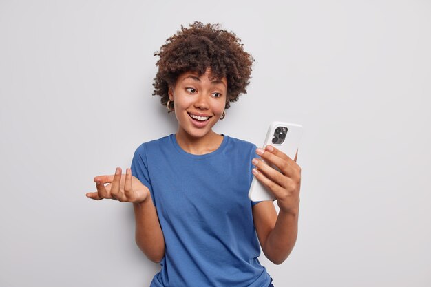 Позитивная миллениальная девушка делает видеозвонок, общается с лучшим другом на расстоянии, держит мобильный телефон, использует бесплатное подключение к Интернету, одетая в повседневную синюю футболку, изолированную на белом фоне
