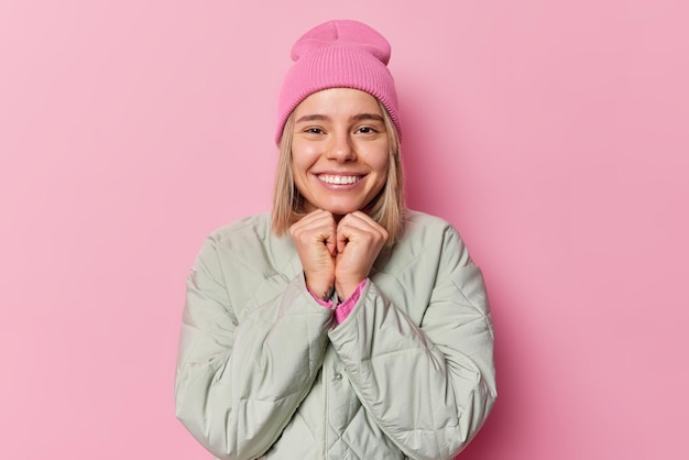 Positive millennial girl smiles broadly keeps hands under chin smiles happily wears hat and jacket feels satisfied listens someone with interest isolated over pink background. Sincere emotions concept