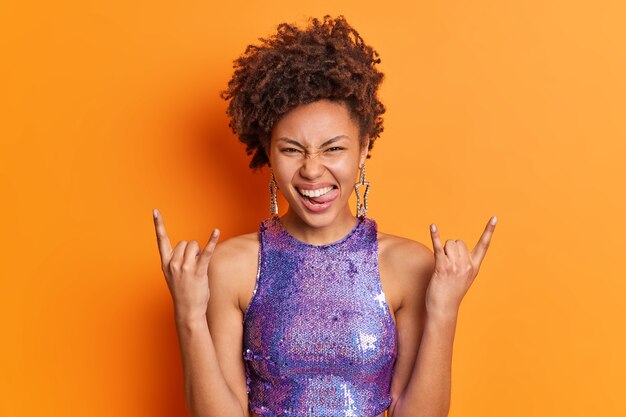 Positive millennial girl with Afro hair sticks out tongue makes rock n roll gesture horns with fingers has fun listens favorite music on party